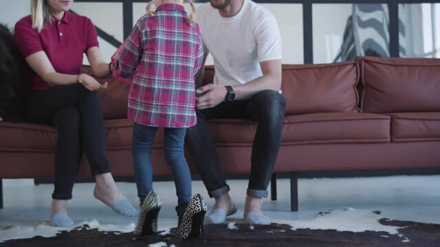 Back view of little Caucasian girl walking in high-heels to sofa. Young smiling man and woman sitting on couch and admiring daughter enjoying weekends at home. Lifestyle, leisure, childhood.