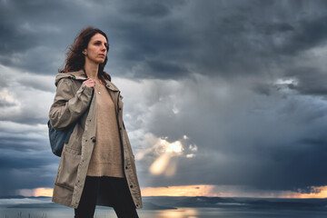 side view of a close up silhouette of a young stylish girl with a backpack on a hilltop with grass and cloudy sunset sky background. Wildlife and outdoor concept.