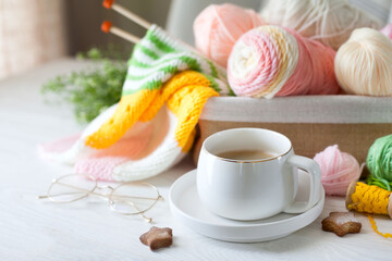 Knitted bright striped fabric. Woolen and cotton yarn lies in a basket. Cappuccino coffee, cookies and glasses lie on a white wooden background.