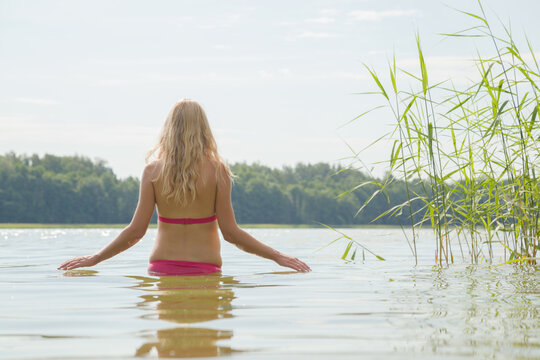 One young woman in pink swimsuit alone slowly going to swim in lake. Warm morning in summer. Peaceful atmosphere. Back view.
