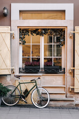 Fototapeta na wymiar Old white wooden window with rectangular frames for glass and christmas garland with branches with a flower pot with flowers and old-fashioned bike near the beige facade.