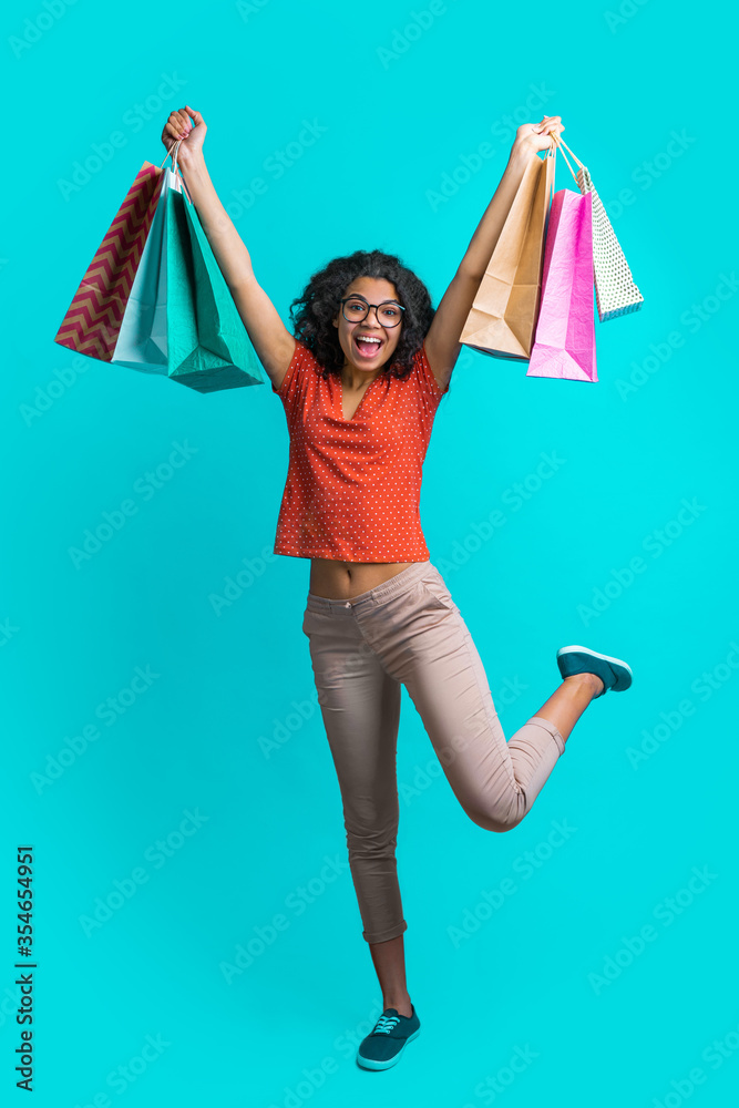 Wall mural Vertical shot of excited dark skinned girl with a bunch of shopping bags jumping over bright blue background with broad smile on her face