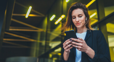 Lifestyle portrait business woman evening city and using mobile gadget, girl banker dressed in business style listening to music wireless earphones and typing text message on smartphone device