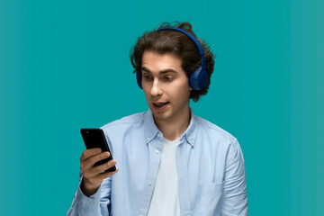 What is it. Portrait of a young beautiful man wearing white t-shirt and blue shirt in blue headphones holding mobile phone in hand and looking at him with wide open eyes
