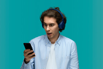Shock sound. Portrait of a young beautiful man wearing white t-shirt and blue shirt in blue headphones holding mobile phone in hand and looking at him with wide open mouth