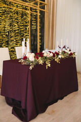 Presidium, banquet table with marsala tablecloth, candles, setting cards, napkins, plates, cutlery, glasses, composition of flowers of marsala rose. Candles, decor in gold, wine and marsala colors