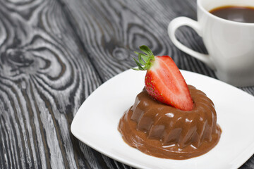 Chocolate fudge garnished with half a strawberry with petals. A cup of coffee. Saucer with dessert stands on brushed pine boards painted in black and white.
