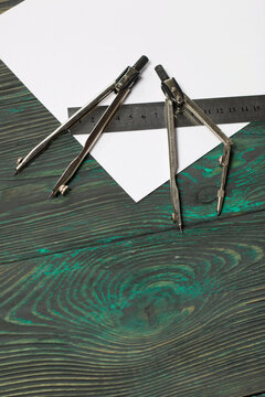 Set for drawing. Two compasses, a metal ruler and a sheet of white paper. They lie on pine boards painted in black and green.