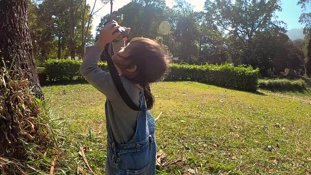 Asian child girl with camera in nature fields. A young girl photographer takes pictures of tree in the park. Concepts of technology and education, learning about nature.