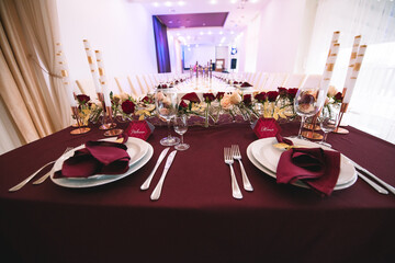 Banquet table with marsala tablecloth, candles, setting cards, napkins, plates, cutlery, glasses, composition of flowers of marsala rose. Caption: 