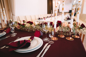 Banquet table with marsala tablecloth, candles, setting cards, napkins, plates, cutlery, glasses, composition of flowers of marsala rose. Candles, decor in gold, wine and marsala colors