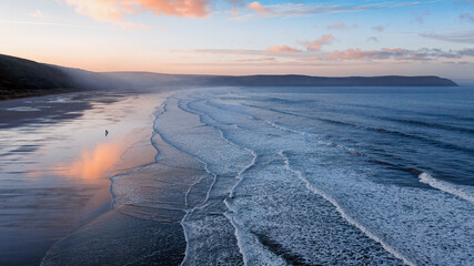 Aerial view of Woolacombe beach  and Baggy Point at dawn, waves breaking on a beach where the sunlit clouds are reflected in the standing water. A man and his dog in silhouette walk towards the sea.