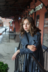 young thoughtful pretty girl in gray coat and turtleneck poses in front of red building. Street style portrait photo session of attractive female, urban photoshoot of beautiful elegant model