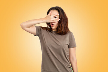 Portrait of a surprised young Caucasian woman, covering her face with her hand, and peeking through her fingers. Yellow background. Human emotions, the concept of facial expression
