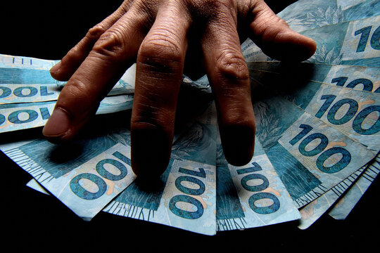 Notas de cem reais brasileiros e m&atilde;o de homem no escurid&atilde;o