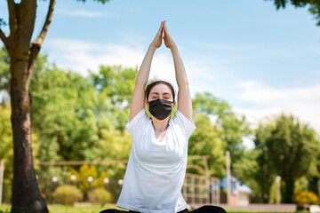 Yoga.A young woman in sports clothing and a medical mask,lying in the Park on the grass and meditating with her hands raised above her head.The concept of healthy lifestyle and protection from viruses