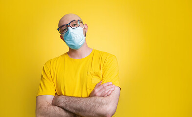 Against a yellow background stands a bald man in a yellow t-shirt, glasses and a medical mask, confidently crossing his arms over his chest. Concept of quarantine, self-isolation and coronavirus