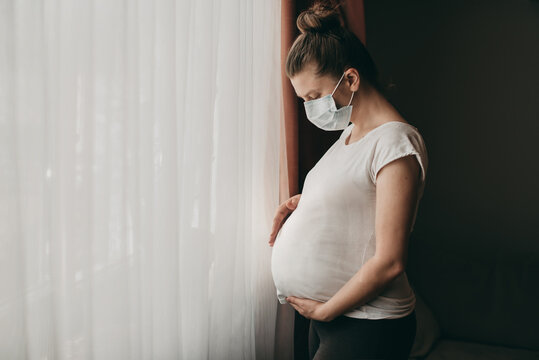 Young beautiful pregnant woman in medical surgical mask and white t-shirt quarantined at home by the window. Coronavirus, virus, isolation, stay at home.