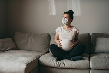 Young beautiful pregnant woman in medical surgical mask and white t-shirt sits on gray couch on quarantine at home. Coronavirus, virus, isolation, stay at home.