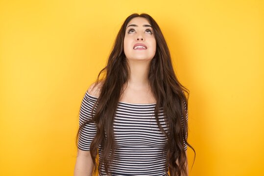 Portrait Of Mysterious Charming Female With Straight Hair Looking Up With Enigmatic Smile. Beautiful Smiling Girl Looking Up Standing Against Gray Wall.