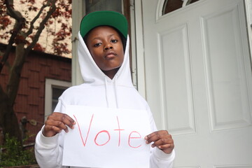 Black Kid holding white paper sign with word Vote written in red house porch background