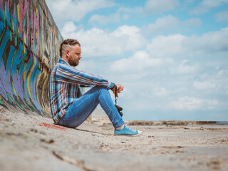 bearded hipster man with mohawk sits on a concrete pier near the sea and looks into the distance against the sky and clouds