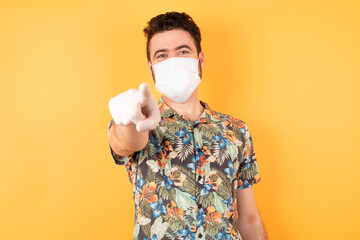 Young caucasian man wearing hawaiian shirt and medical mask pointing at camera with a satisfied, confident, friendly smile, choosing you against flat yellow background