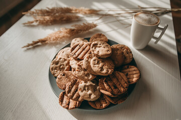 Homemade baked cookies on a plate on
white wooden table and cup of coffee, dried grass, dried flowers and sunlight, breakfast in the morning. To cook delicious quarantine at home. Rustic style.