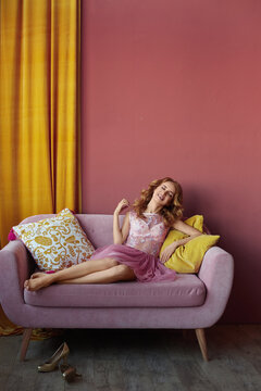 Young Happy Woman In A Pink Cocktail Dress Took Off Her Shoes And Rests On The Couch In The Home Interior. Concept Of Summer Fashion. Tired Slim Girl Has A Rest