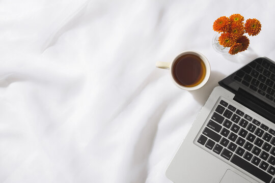 Top view of morning tea in voile fabric background with a laptop, mug of tea and a vase of orange flowers with space for text.