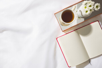 Top view of morning scene in voile fabric background with a red notebook, mug of tea and a vase of white flowers in a mirrored brass tray, with space for text.
