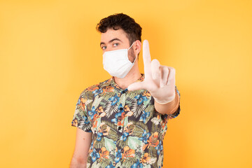 Young handsome businessman wearing hawaiian shirt and medical mask standing over yellow isolated background making fun of people with fingers on forehead doing loser gesture mocking and insulting.