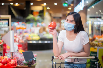 An Asian woman wearing a surgical mask is walking in a supermarket. She is choosing fruit. She is smiling under the mask. This image has a copy space.