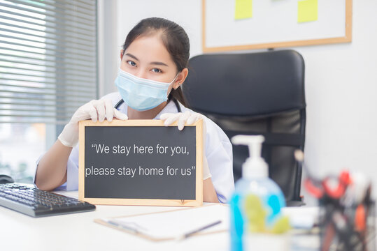 An Asian Female Doctor Wearing A Surgical Mask Sat In The Hospital Office. She Holds A Board That Says 