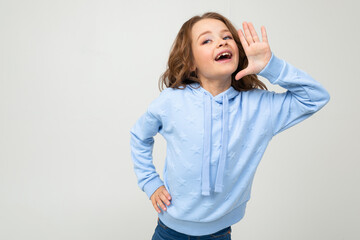 European charming girl in a blue hoodie tells the news while holding hands at the mouth on a light background