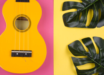 Body of a bright yellow ukulele guitar and monstera leaves on a split pink and yellow background (as a tropical vacation concept)