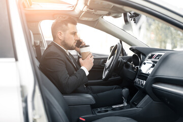 Handsome caucasian businessman driver inside in vehicle  with coffee and gadget and check mail and talking with partners