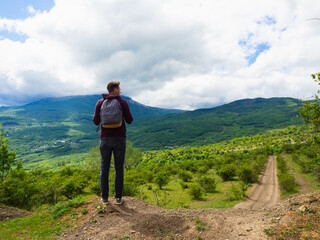 Fototapeta premium A young guy in a red hoodie and gray backpack travels in the mountains among green trees and clouds.