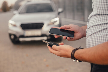 Young handsome man with smartphone and car keys drink black coffee and wait for friend near city parking