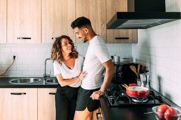 Young couple of happy lovers look at each other in the kitchen of their house. The woman looks happily and smiling at her bearded man. They both wear comfortable clothes from being at home.