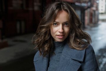 young happy smiling pretty girl in gray coat and turtleneck poses in front of building. Street style portrait photo session of attractive female, urban photoshoot of beautiful elegant model