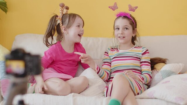 Shooting a Video Blog for Children with Camera on Tripod. Production of Kid's Content. Two Cute Cheerful Little Caucasian Girl Talking and Laughing, Sitting on a Couch. Cozy Interior with Yellow Wall.
