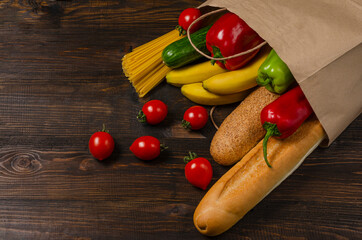 Paper grocery bag with long loafs, spaghetti, bananas, cucumber, hot and sweet peppers. Quarantine food. Products on dark brown wooden wooden. Donation of food during quarantine. Home delivery