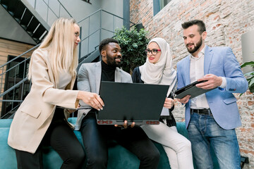 Team of multiethnic smart male and female business people leaning on the blue sofa in modern office, brainstorming and woring together, using laptop and tablet. Business and digital technologies