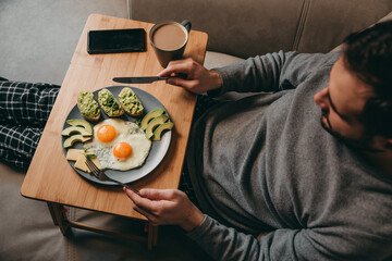 The guy sits on the couch with breakfast table and eats breakfast on a plate of fried eggs, bread, avocado toast, cheese and coffee, a smartphone on the table.