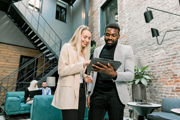 Two young business colleagues, Caucasian woman and African man, working together in the office using the computer tablet and brainstorming. Colleagues on the background