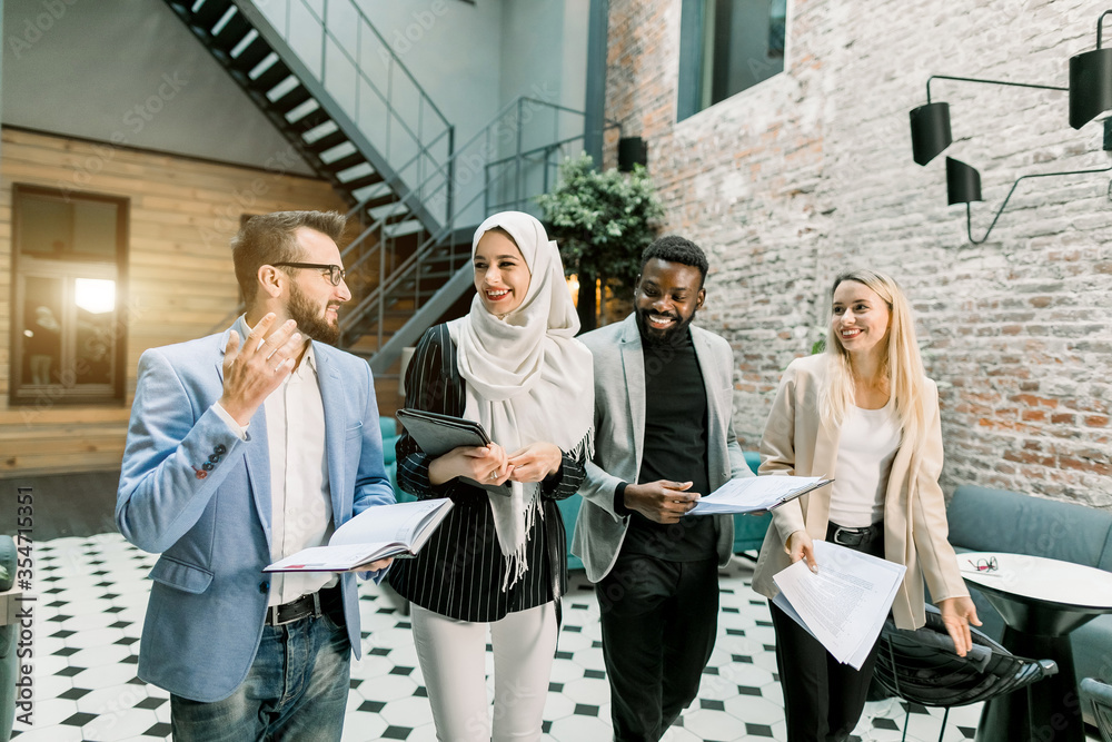 Wall mural Business multiethnical team. Happy young multiethnic businessmen and businesswomen walking through the modern office room, holding tablet and papers, after their work day