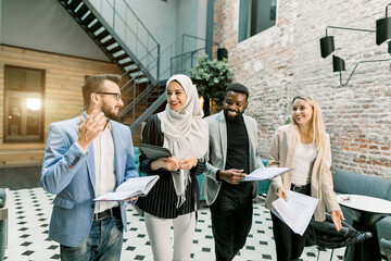 Business multiethnical team. Happy young multiethnic businessmen and businesswomen walking through the modern office room, holding tablet and papers, after their work day