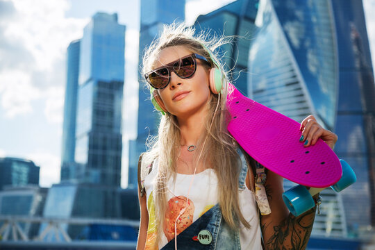 Summer lifestyle image of trendy pretty girl with pink pennyboard on skyscrapers background. Tattoo model. Smiling and listening music in stylish headphones. Wearing stylish sunglasses, denim overalls