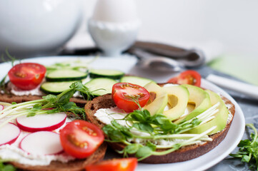 Vegetarian sandwiches with cream cheese, cucumber, avocado, garden radish, tomatoes and peas sprouts on white plate. Egg mist on white stand. Breakfast with vegetarian food.
